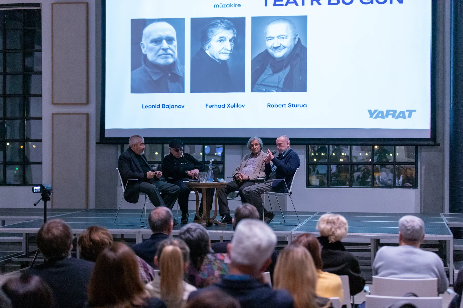 The speakers on stage beneath a projection naming Leonid Bazhanov, Farhad Khalilov and Robert Sturua.