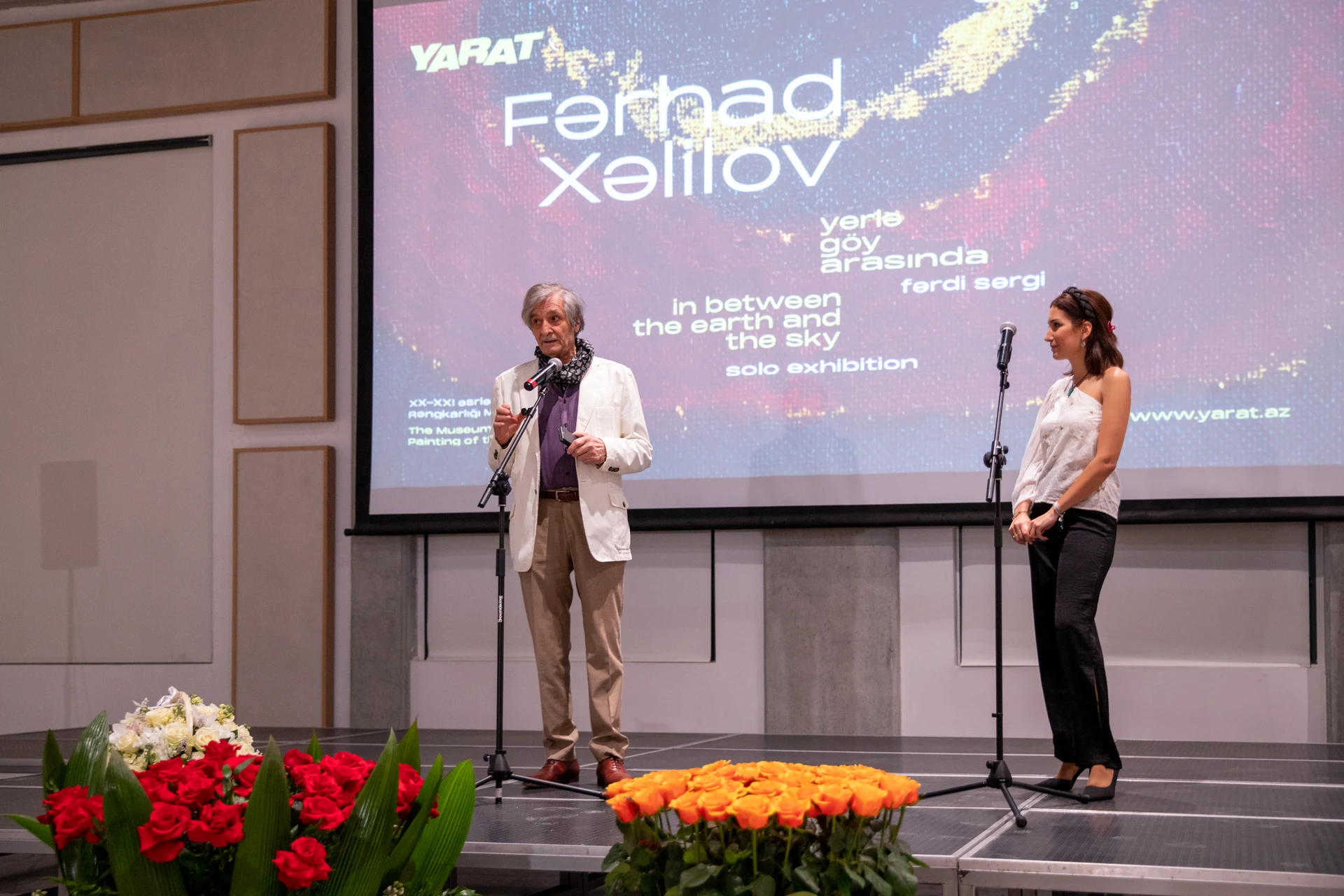 Farhad Khalilov and a presenter address the opening audience in front of the exhibition title projection.